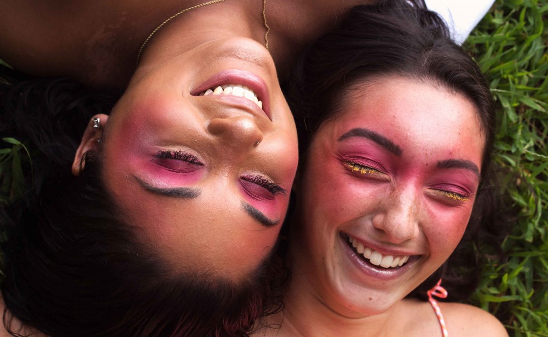 close-up of two people with colorful makeup, laughing and laying in the grass